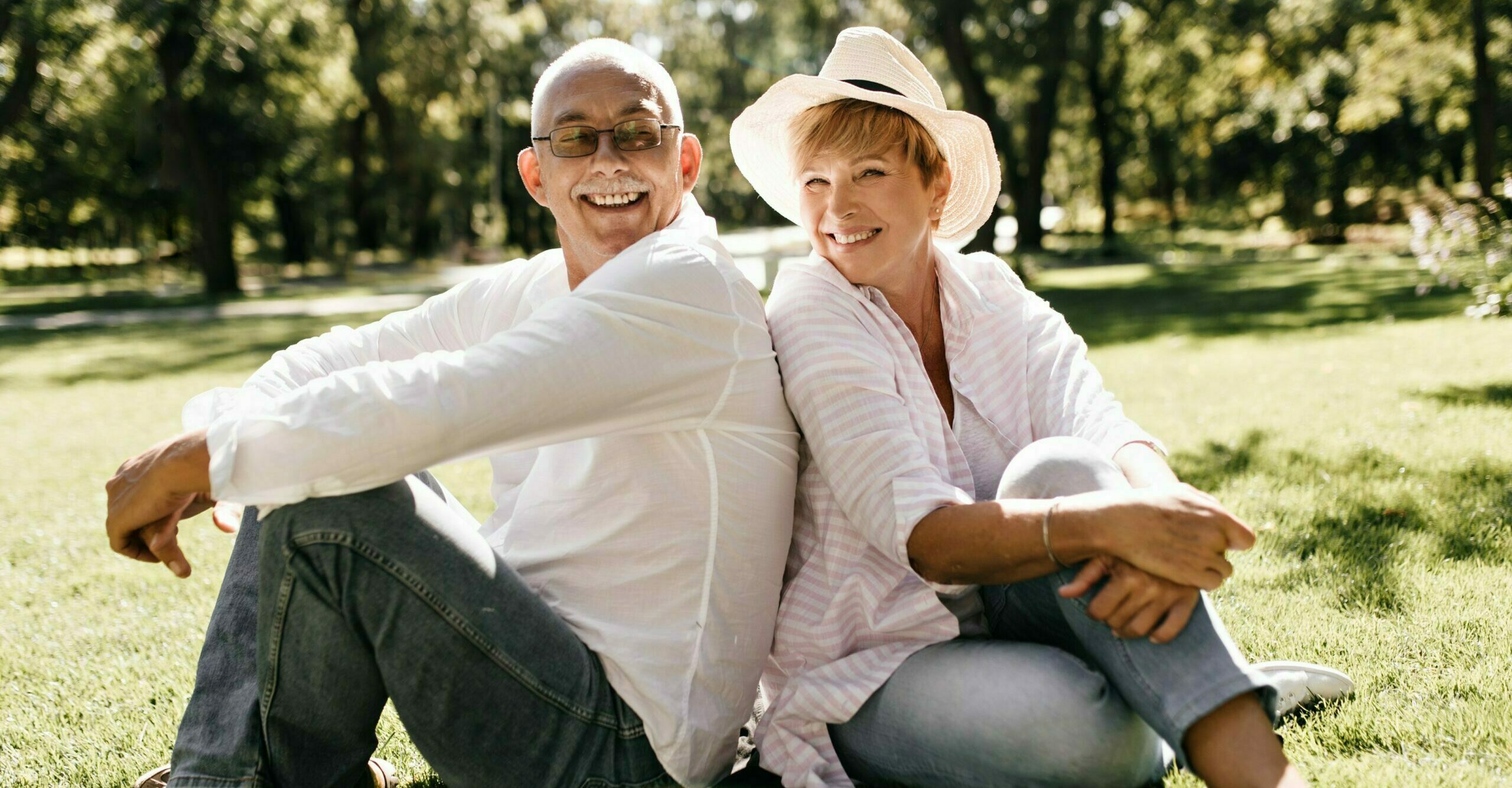 Positive lady with cool hat in striped stylish blouse and jeans smiling and sitting on grass with man in eyeglasses and light shirt outdoor..